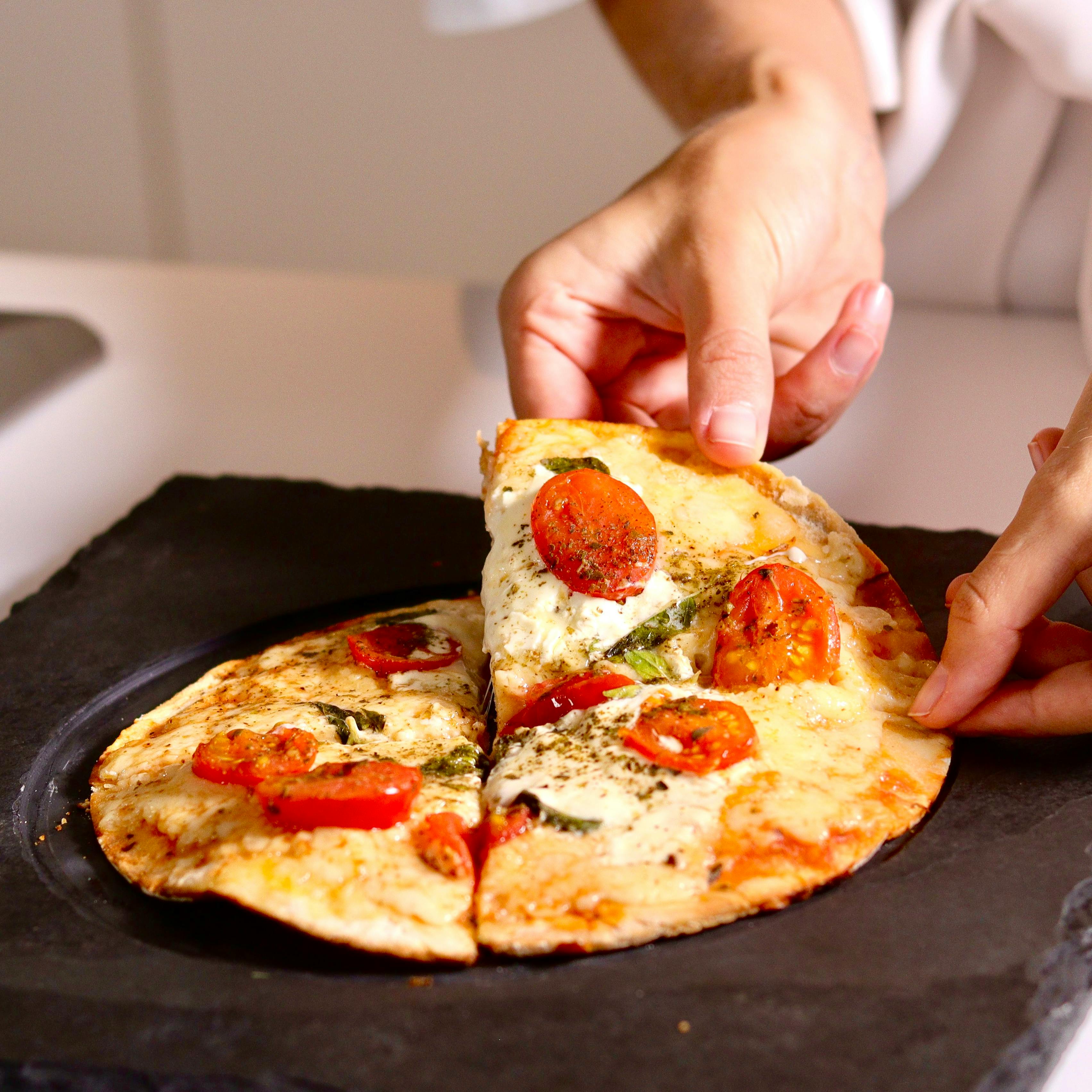 A hand picking up a slice of cooked margherita pizza from a black square plate